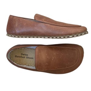 Pode incluir: Um par de mocassins de couro castanho com uma costura ao redor da sola. Os mocassins têm um design simples e são feitos pela Swiss Barefoot Shoes.