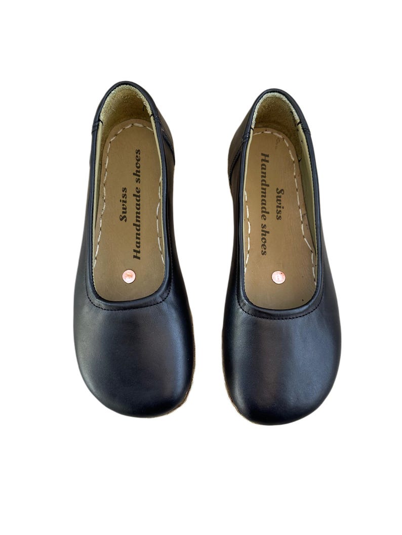 May include: A pair of black leather ballet flats. The shoes have a rounded toe and a stitched sole. The inner sole is tan with the text "Swiss Handmade shoes".