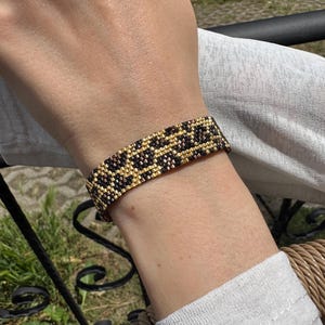 May include: A woven beaded bracelet featuring a repeating leopard print pattern in metallic gold, bronze, and black glass seed beads. The cuff-style bracelet has a flat, rectangular shape and is constructed with a tight, uniform grid of small beads.