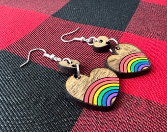 Puede incluir: Un par de pendientes de madera en forma de corazón con un diseño de arcoíris. Los pendientes están sobre un fondo a cuadros rojo y negro.