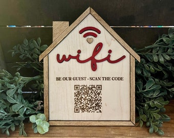 May include: Wooden house-shaped sign with the word "wifi" in red cursive lettering and a wifi symbol above it. The sign also includes the text "BE OUR GUEST - SCAN THE CODE" and a black and white QR code.
