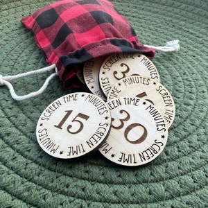 Op de afbeelding: Een set houten schermtijd-timertokens in een rood-zwart geruite tas. De tokens zijn rond en hebben de tekst "Schermtijd" en "Minuten" erop gedrukt. De nummers 3, 15 en 30 zijn zichtbaar op de tokens.