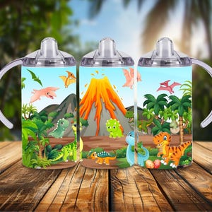 Puede incluir: Tres vasos con boquilla con ilustraciones de dinosaurios. Los vasos tienen tapas y asas transparentes. Los diseños de dinosaurios incluyen un volcán, palmeras y varios dinosaurios de dibujos animados en tonos verdes, naranjas y azules.