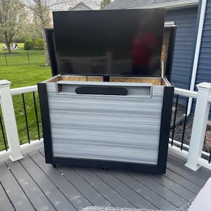 Puede incluir: Un mueble de entretenimiento para exteriores con un televisor negro grande. El mueble es gris y negro con una barra de sonido. La unidad está sobre ruedas y se encuentra en una terraza gris.