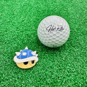 May include: A blue and yellow golf ball pin with a white shell and the text "High Roller" on a white golf ball.