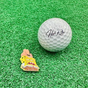 May include: A white golf ball with the text "High Roller" in black script sits on green turf next to a yellow and brown cartoon character pin wearing a cowboy hat. The pin is a golf accessory.