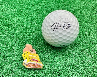 May include: A white golf ball with the text "High Roller" in black script sits on green turf next to a yellow and brown cartoon character pin wearing a cowboy hat. The pin is a golf accessory.