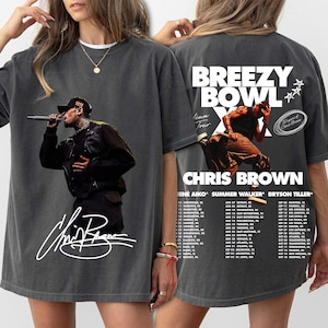 May include: Dark gray t-shirt featuring a concert image of Chris Brown. The front displays a photo of the artist with his signature. The back of the shirt has the text "Breezy Bowl" and a tour schedule.