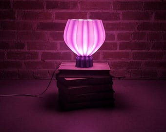 May include: A glowing, pink table lamp with a ribbed shade sits atop a stack of books. The lamp's base is a darker shade of purple. The background features a brick wall, all illuminated with a pink hue.