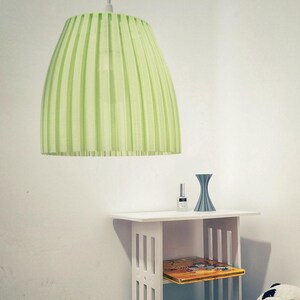Può includere: Un paralume plissettato verde chiaro pende da un apparecchio a soffitto bianco. Il paralume è realizzato in un materiale translucido e ha una superficie leggermente strutturata. Un tavolino bianco con uno scaffale è in primo piano. Un piccolo vaso blu e una bottiglia di smalto per unghie sono sul tavolo.