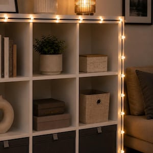 Könnte beinhalten: Ein weißes Regal mit mehreren Fächern, das Pflanzen, Boxen und eine Lampe enthält. Eine Lichterkette mit kleinen, warmweißen Lichtern ist am Rand und an der Seite des Regals angebracht und erzeugt ein sanftes Leuchten.