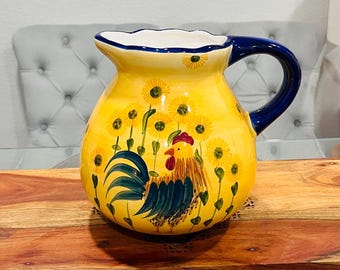 Puede incluir: Una jarra de cerámica amarilla con asa azul y un diseño de gallo. El gallo está pintado en tonos de verde, amarillo, rojo y marrón. La jarra está decorada con flores amarillas.