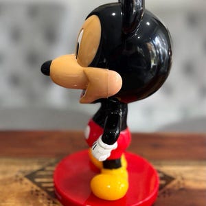 K&ouml;nnte beinhalten: Eine schwarze und rote Mickey Mouse Figur mit gelbem Sockel. Die Figur steht auf einem roten Sockel mit einem schwarz-wei&szlig;en Muster.