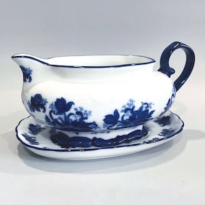 May include: White porcelain gravy boat with a matching saucer. The set features a blue floral pattern and a dark blue handle. The rim of the gravy boat and saucer are also trimmed in dark blue. A classic design for serving sauces and gravies.