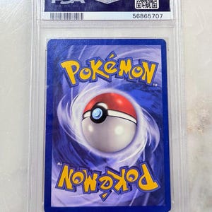 May include: A Pok&eacute;mon trading card encased in a clear plastic holder. The card features a blue background with a Pok&eacute; Ball design and the word "Pok&eacute;mon" in yellow. The holder has "PSA" branding and a serial number.