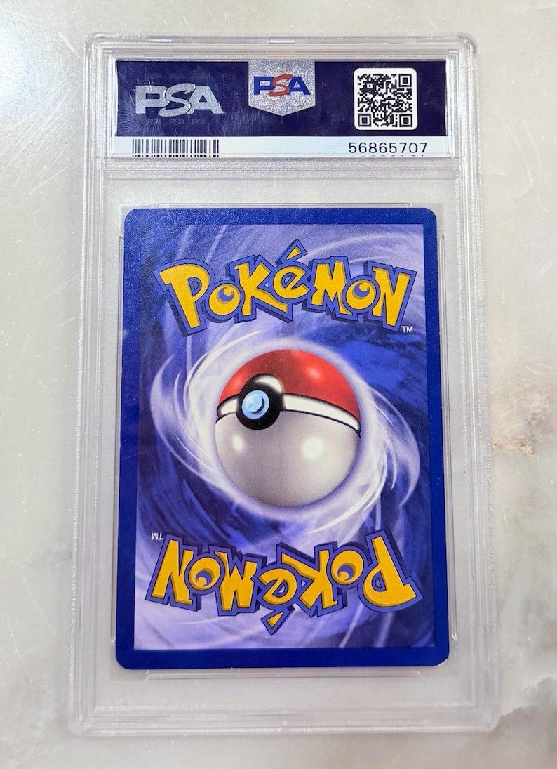 May include: A Pok&eacute;mon trading card encased in a clear plastic holder. The card features a blue background with a Pok&eacute; Ball design and the word "Pok&eacute;mon" in yellow. The holder has "PSA" branding and a serial number.