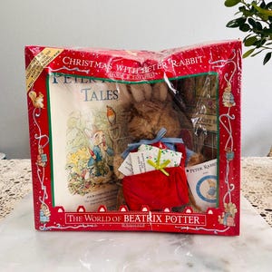 Puede incluir: Un set de regalo navideño con Peter Rabbit. La caja roja contiene un peluche, un libro titulado "Peter Rabbit Tales" y un calcetín rojo. La caja está decorada con ilustraciones de Peter Rabbit y otros personajes.