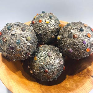 May include: Four ornate, silver-colored spheres with intricate floral patterns and embedded colorful gemstones. The spheres are displayed in a natural wooden bowl, showcasing a blend of textures and colors.