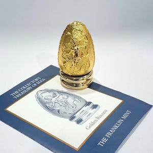 May include: A golden egg sculpture with intricate relief details, displayed on a gold and silver stand. The egg is positioned on a blue and white card with the text "THE COLLECTOR'S TREASURY OF EGGS" and "Golden Rococo".