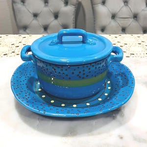 May include: A bright blue ceramic soup bowl with a matching lid and saucer. The bowl has two handles and a handle on the lid. The bowl and saucer are decorated with a dotted pattern and stripes.