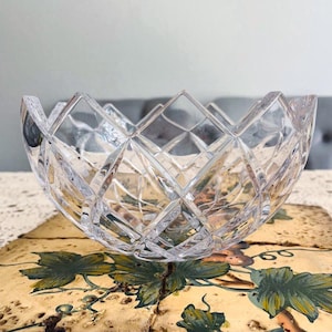 Könnte beinhalten: Eine klare, facettierte Kristallschale mit einem Rautenmuster. Die Schale hat einen gewellten Rand und steht auf einer dekorativen Oberfläche mit grünen und braunen floralen Designs. Diese elegante Schale eignet sich zum Servieren oder Ausstellen.