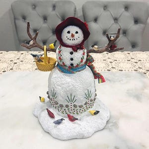 Op de afbeelding: Een decoratief sneeuwpopfiguur met een rode hoed, sjaal en bloemaccenten. De sneeuwpop heeft een takachtige arm met vogels erop en een klein mandje. Verschillende vogels zijn verspreid rond de basis, die ontworpen is om op sneeuw te lijken.