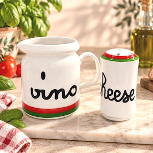 May include: A white ceramic pitcher with the word vino in black script and a red and green base stripe, paired with a white ceramic shaker labeled cheese in black script with a red and green lid featuring small black dots.