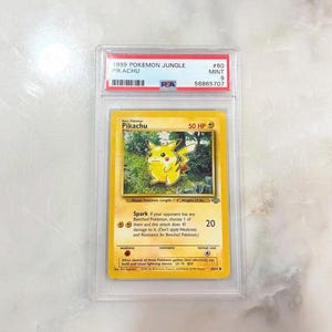 May include: A graded Pok&eacute;mon card featuring Pikachu from the 1999 Jungle set. The card is yellow with a Pikachu illustration and text. The card is encased in a clear protective holder with a label indicating its condition and serial number.