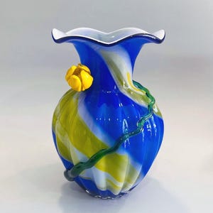 May include: A decorative glass vase with a ruffled white rim. The vase features a vibrant design of blue, yellow, and white swirls, with a yellow flower and green vine accent. The vase is a unique piece of art.