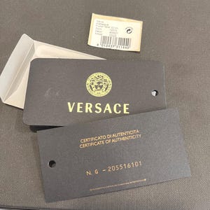 May include: Black Versace product tags with gold lettering and a gold Medusa head logo. One tag reads "CERTIFICATO DI AUTENTICIT&Agrave; CERTIFICATE OF AUTHENTICITY". A small white tag with product information is also present.