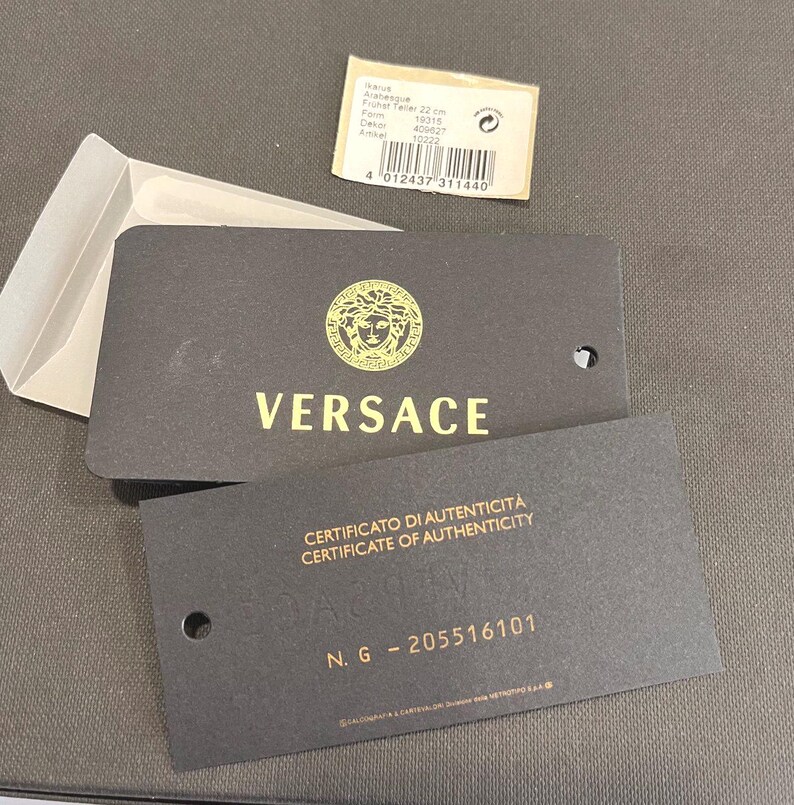 May include: Black Versace product tags with gold lettering and a gold Medusa head logo. One tag reads "CERTIFICATO DI AUTENTICIT&Agrave; CERTIFICATE OF AUTHENTICITY". A small white tag with product information is also present.
