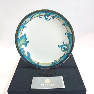 May include: White ceramic plate with a teal and gold decorative border and teal and green floral design. The plate is on a black box with a Versace logo on a card.