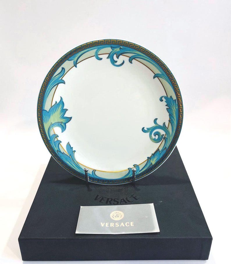 May include: White ceramic plate with a teal and gold decorative border and teal and green floral design. The plate is on a black box with a Versace logo on a card.