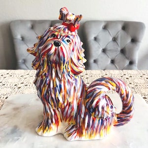Könnte beinhalten: Bunte Keramik-Hundefigur mit Schleife auf dem Kopf. Der Hund ist mit vertikalen Strichen in Rot, Blau, Gelb und Weiß bemalt. Die Figur hat einen lockigen Schwanz und sitzt aufrecht.