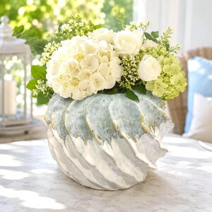 May include: A ceramic vase with a spiraled, shell-like texture featuring a white base and a textured, seafoam green upper section with gold-toned accents. The vase is filled with a large arrangement of white hydrangeas, white roses, and green foliage.