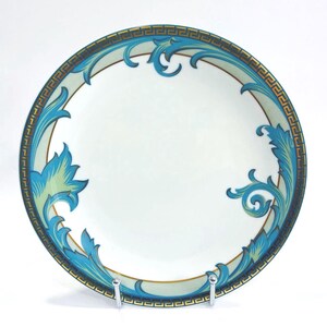 May include: A white ceramic plate with a decorative border and ornate blue and gold leaf design. The plate features a Greek key pattern around the rim and is supported by a silver stand.