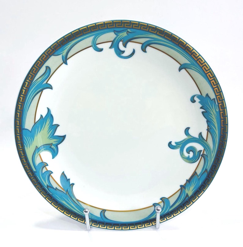May include: A white ceramic plate with a decorative border and ornate blue and gold leaf design. The plate features a Greek key pattern around the rim and is supported by a silver stand.