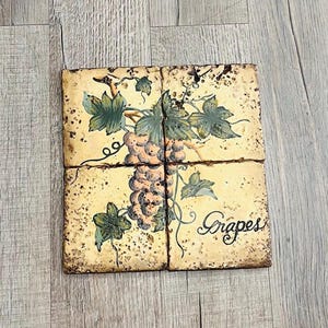 May include: Four square tiles arranged together, featuring a hand-painted design of grapes with green leaves and the word "Grapes" in script. The tiles have a distressed, aged appearance with a beige background.