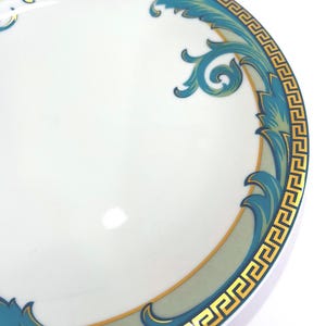 May include: White ceramic plate with a teal and gold decorative border. The border features a Greek key design and stylized floral elements. The plate has a glossy finish and is set against a white background.