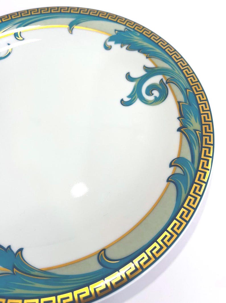 May include: White ceramic plate with a teal and gold decorative border. The border features a Greek key design and stylized floral elements. The plate has a glossy finish and is set against a white background.