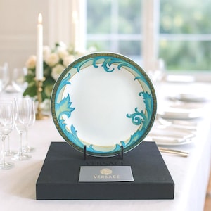 May include: A white ceramic plate with a teal and gold decorative border and accents, displayed on a black stand. The plate features a detailed design with floral and scroll motifs. A card with the Versace logo is in front of the plate.