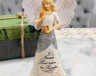 May include: A decorative angel figurine with blonde hair, holding orange roses. The angel wears a white dress with a silver patterned skirt and wings. The text on the dress reads "A Nana's Love makes life Sweeter."