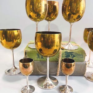 Puede incluir: Una colección de copas de vino doradas y copas de licor más pequeñas. Las copas tienen una superficie brillante y reflectante y están dispuestas sobre una superficie blanca. Los tallos y las bases son de color plateado.
