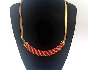 Puede incluir: Un collar dorado con una pieza central roja y curvada. La pieza central está envuelta con alambre dorado. El collar se exhibe en una cabeza de maniquí negra con un diseño.
