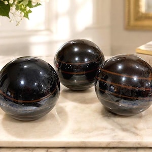 Puede incluir: Tres objetos decorativos esféricos con un acabado negro pulido y sutiles bandas horizontales de color marrón. Cada esfera presenta una superficie lisa y reflectante que capta la luz, creando un aspecto brillante en las formas redondeadas.
