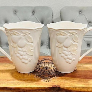 May include: Two white ceramic mugs with a pear and grape design. The mugs have a raised design and are on a wooden surface.