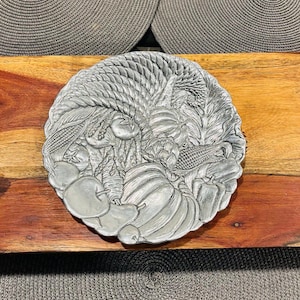 Puede incluir: Una bandeja de servir de metal plateado con un diseño en relieve de diversas frutas y verduras. La bandeja es redonda y tiene un estilo rústico, de granja.