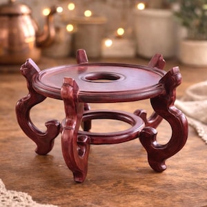 May include: A dark red, wooden decorative stand with a round top and a central hole. The stand has three curved legs and a circular base. The stand is designed to hold a small object. The background includes a copper teapot and soft lighting.
