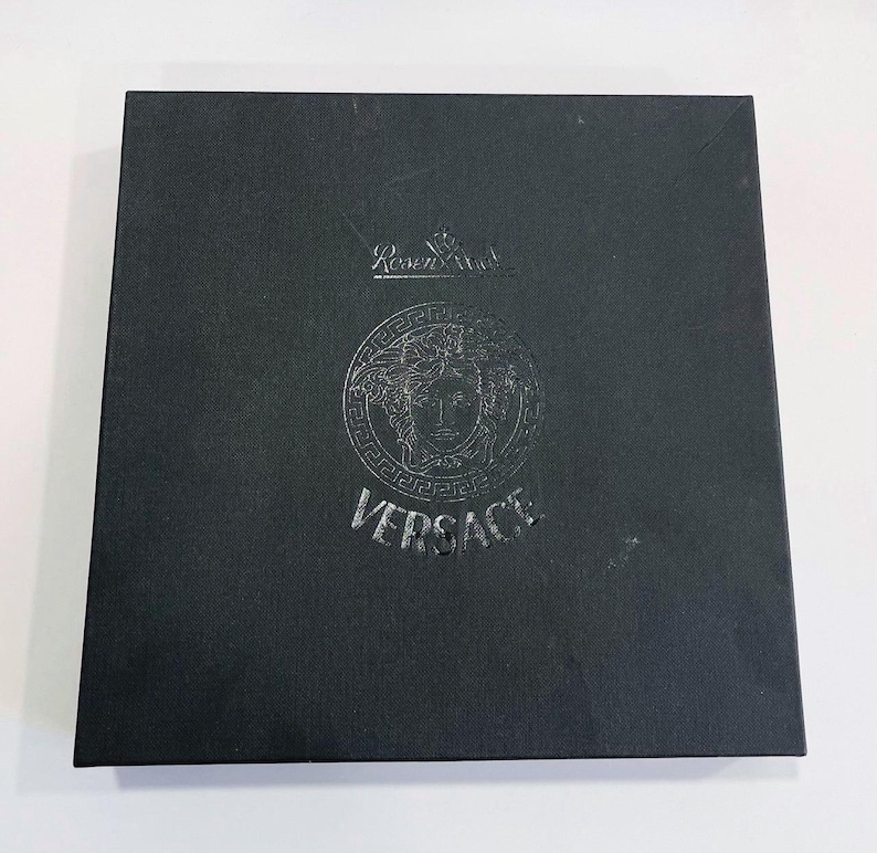May include: A black square box with the Versace logo embossed on the front. The logo features a Medusa head and the word "VERSACE" below it. The box also has the "Rosenthal" logo at the top.