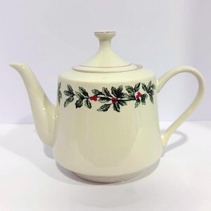 Może przedstawiać: Kremowy ceramiczny imbryk z zakrzywionym dziobkiem i uchwytem. Imbryk posiada dekoracyjną girlandę z zielonych liści i czerwonych jagód wokół korpusu. Pokrywka ma mały uchwyt i złote obramowanie.